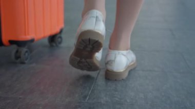 Yakından bakınca, havaalanında elinde turuncu bir bavulla yürüyen bir kadın görülüyor. Adımlarına, ayakkabılarına ve ileri hareket hareketlerine odaklanıyor..