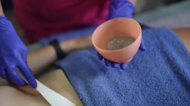 Estetisyen tarafından hazırlanan abdominal şekerleme macunu uygulaması. Kadın kalçasında havluyla yatarken, terapist mor eldivenler giyiyor.