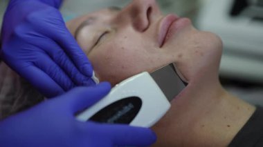 Modern bir güzellik salonunda derin bir yüz temizleme prosedürü için ultrasonik cilt temizleyici kullanan mavi eldivenli profesyonel kozmetolog.