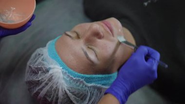 Mor eldivenli kozmetolog, güzellik salonundaki fırçayla rahatlatıcı bir kadının yüzüne şeffaf gençleştirici jel sürüyor.