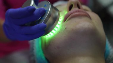Kafkasyalı genç bir kadın ışık terapisi görüyor. Bir kozmetikçi, yüz tedavisinde profesyonel bir cihaz kullanıyor.