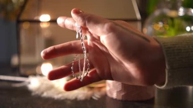 Parıldayan gümüş bir kolyeyi nazikçe tutan dövmeli birinin yakın çekimi. Sıcak, yumuşak ışık narin mücevherleri aydınlatır.