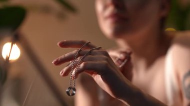 Omzunda dövmesi olan genç bir kadın hafifçe aydınlatılmış bir odada gümüş bir kolyeyi nazikçe tutuyor ve inceliyor.
