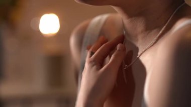 Genç bir kadının boynunu ve ellerini sıcacık aydınlatılmış bir odada gümüş bir kolye takarken yakın plan. Hazırlanıyor ve hazırlanıyor.