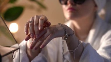 Bornozlu ve güneş gözlüklü bir kadının gümüş zincirli bilezik denemesi. Güzel bir manikürle elleri odağında.