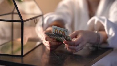 Bir kadının 100 dolarlık banknotları sayarken çekilmiş yakın plan görüntüsü. Beyaz bornoz giyen kişi parayı karanlık bir masaya koyar.