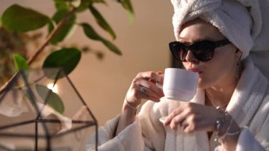 Beyaz bornozlu ve kafasında havluyla sabah güneşinin tadını çıkaran genç bir kadın. Nazikçe kahve içiyor ve kameraya bakıyor.