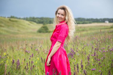 Parlak fuşya rengi elbiseli sarışın kadın yazın elinde çiçeklerle tarlada yürür.