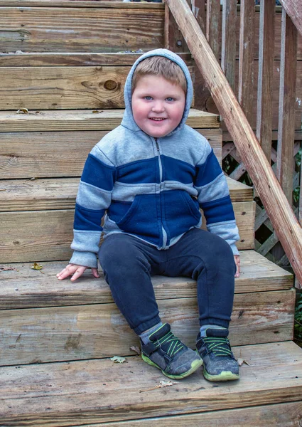 一个快乐的小男孩穿着蒙面夹克 面带微笑地坐在木板台阶上 — 图库照片