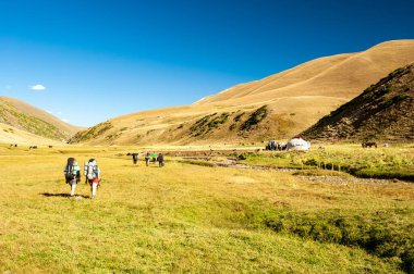Bir grup yürüyüşçü dağ manzaralı kayalık geçitten yurt yerleşimine doğru iniyor. Sary Chelek Gölü, Sary-Chelek Celal Abad bölgesi, Kırgızistan, Orta Asya 'da yürüyüş yapan genç erkek ve kadınlar.