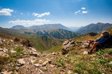 Young trekker man resting in rocky pass with mountain top views in high mountains. Young men and women hiking near Sary Chelek lake, Sary-Chelek Jalal Abad region, Kyrgyzstan, Trekking in Central Asia.