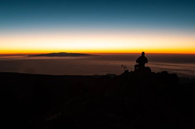 El Teide Ulusal Parkı 'ndaki Pico del Teide Dağı' nda şafak vakti oturan ve güneşin doğmasını bekleyen farlı bir turist. Tenerife, Kanarya Adaları, İspanya