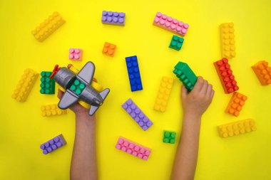 Çocukların elleri sarı arka planda tasarımcı parçalarından yapılmış bir uçağı tutuyor. Çok renkli inşaat blokları. Çocukların eğitici oyunu. Arkaplan ve doku.