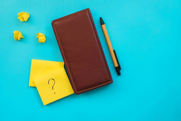 Stickers in the form of a yellow square and a leather notebook lie on a blue background. Notebooks for notes and reminders. A question mark is written on the sheet.Next to the notebook is a pen.