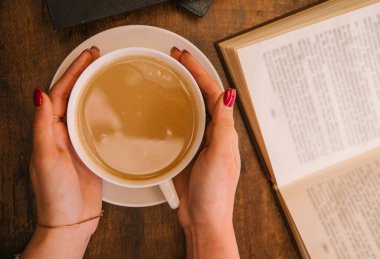 Kadın eli bir bardak kapuçino tutuyor. Yanındaki ahşap masada kitaplar var. Samimi bir kafe. Faydalı bir boş zaman. Eğitim ve kendi kendini geliştirme fikri.