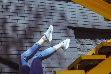 Dişi bacaklar tuğla bir duvarın arka planına ve merdivenlere dayanıyor. Kotlu bacaklar ve spor ayakkabıları. Bir fotoğraf için alışılmadık bir poz. Duvara sert gölgeler düşer. 2021 'in trend rengi.