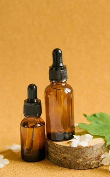 Bir tahta parçasının üzerinde iki şişe kozmetik yağ standı. Yakınlarda yeşil yapraklar ve küçük beyaz çiçekler var. Cilt ve saç bakımı. Alternatif tıp ve aromaterapi..