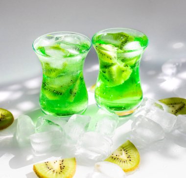 Buz ve kivi dilimli yeşil limonata. Masanın üzerine iki cam bardak ve güneş ışınları düşer. Güneşin güzel gölgesi ve parıltısı. Yaz ferahlatıcı soğuk içecek.