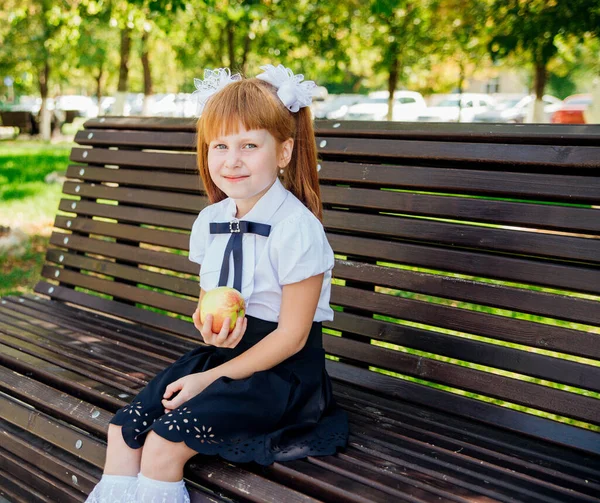 Back to school. A cute little schoolgirl is sitting on a bench in the school yard and holding a green apple in her hands. Proper school meals for lunch. A little girl is going to the first grade.