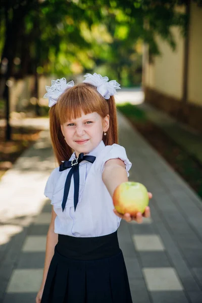 Back to school. A cute little schoolgirl is standing in the park or in the school yard and holding a green apple in her hands. Proper school meals for lunch. A little girl is going to the first grade.