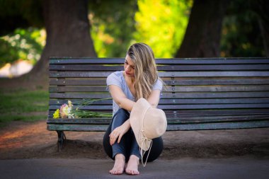 Bir buket çiçekle bankta oturan ciddi, düşünceli ve üzgün yüzlü genç sarışın kadın.