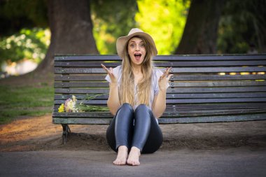 Bir buket çiçekle bankta oturan şaşırmış yüzlü genç sarışın kadın.
