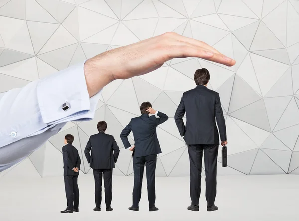 Cuatro hombres de negocios de diferente tamaño dispuestos por la altura de pie bajo una mano enorme. Fondo geométrico gris. Concepto de crecimiento profesional . — Foto de Stock