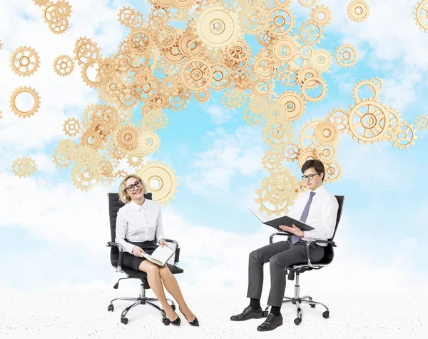 A smiling woman and a man sitting on castor chairs with books. Blue sky and golden cogwheels coming from them at the background. — Stock Photo, Image