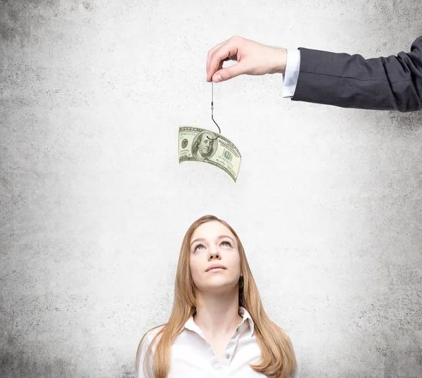 A hand holding a one-hundred dollar banknote on a thread, a young businesswoman looking up at it. Front view. Concrete background. Concept of motivation. — Φωτογραφία Αρχείου