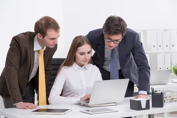 Tres empresarios en una computadora portátil, mujer sentada, fondo de oficina . — Foto de Stock