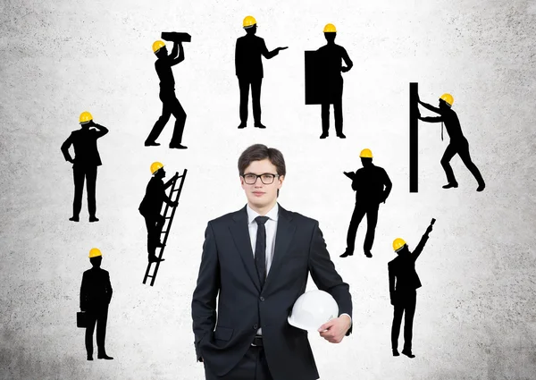 Empresario con casco blanco, siluetas de hombres de negocios con diferentes tareas de construcción a su alrededor, Fondo de hormigón. Concepto de construcción empresarial . —  Fotos de Stock