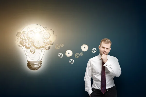 Businessman with hand to chin, light bulb with gears painted aside. Front view. Dark blue background. Concept of new idea. — Stock Photo, Image