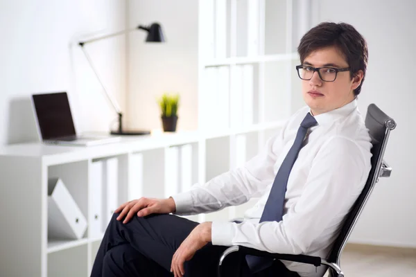 Young businessman wih hand on knees sitting on chair.  Office at background. Concept of work. — Stock fotografie