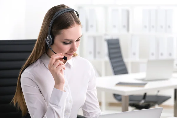 Ung affärskvinna i hörlurar med mikrofon sittande vid datorn. Bakgrundskontor. Begreppet arbete. — Stockfoto