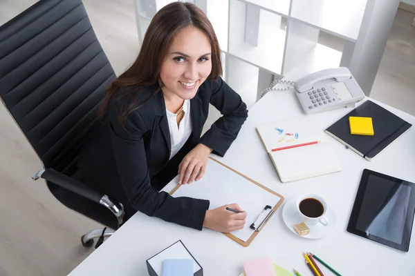 Affärskvinna sitter vid bordet och göra anteckningar. Anteckningsbok, telefon och kopp framför henne. Office. Begreppet arbete. — Stockfoto