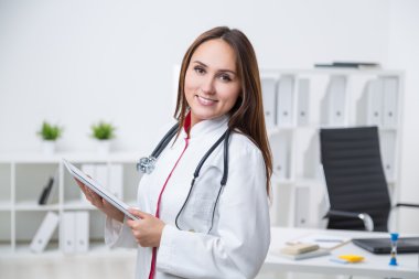 Kadın doktor masada duruyor ve elinde tablet tutuyor. Ön görüş. Arka planda ofis. İş kavramı.