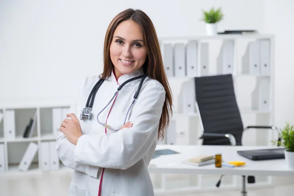 Χαμογελαστή γυναίκα γιατρός στέκεται στο τραπέζι με σταυρωμένα τα χέρια. Μπροστινή όψη. Γραφείο στο παρασκήνιο. Έννοια της εργασίας. — Φωτογραφία Αρχείου