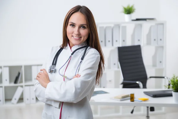 Una doctora sonriente parada en la mesa con las manos cruzadas. Vista frontal. Oficina en segundo plano. Concepto de trabajo . — Foto de Stock