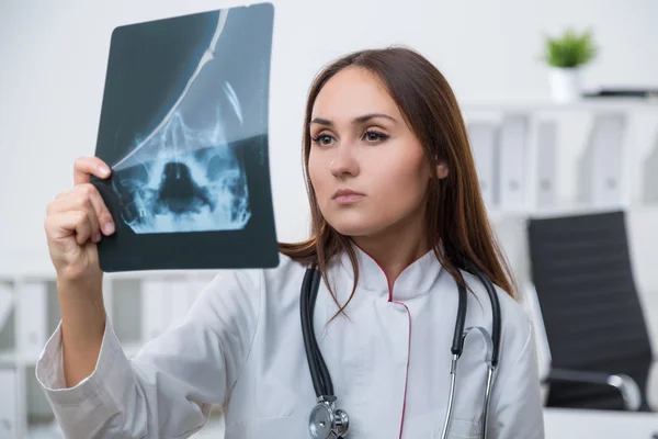 Female doctor standing at table and looking at X-rays, concentrated. Light background. Concept of work. — Stock Photo, Image