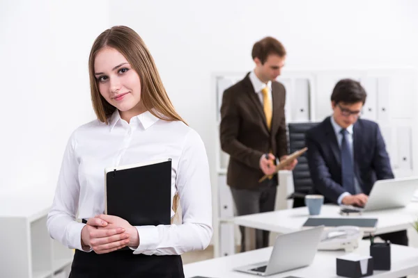 Jovem empresária sorridente com caderno em pé à mesa, dois homens de negócios conversando no fundo. Escritório. Conceito de trabalho . — Fotografia de Stock