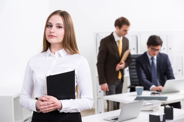 Am Tisch steht eine junge Geschäftsfrau mit Notizbuch, im Hintergrund unterhalten sich zwei Geschäftsleute. Arbeitsbegriff. — Stockfoto