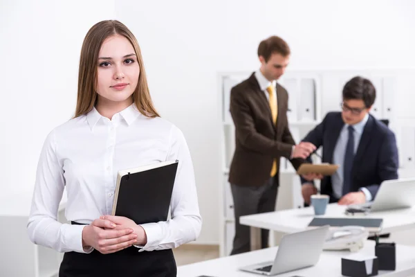 Am Tisch steht eine junge Geschäftsfrau mit Notizbuch, im Hintergrund unterhalten sich zwei Geschäftsleute. Büro. Arbeitsbegriff. — Stockfoto
