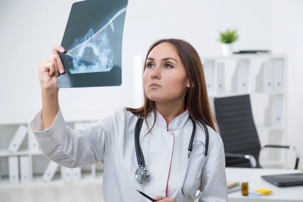 Female doctor standing at table and looking at X-rays. Light background. Concept of work. — Stock Photo, Image