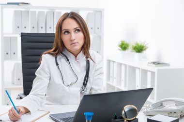 Bilgisayar başında çalışan ve notlar yapan, yukarı bakan kadın doktor. Arka planda ofis. İş kavramı.