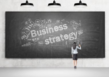 Young businesswoman writing on blackboard, business icons on it, three lamps above. Back view. Concept of business strategy.