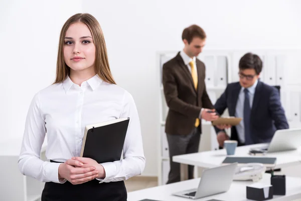 Eine ernsthafte junge Geschäftsfrau mit Notizbuch steht am Tisch, zwei Geschäftsleute unterhalten sich im Hintergrund. Büro. Arbeitsbegriff. — Stockfoto