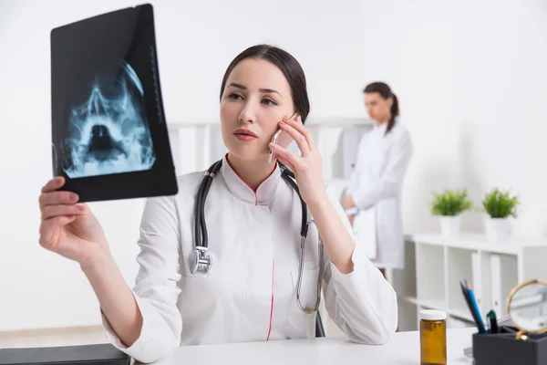 Doctor looking at X-ray of skull and speaking on phone, nurse at background. Concept of medical cosultation. — Stock Photo, Image