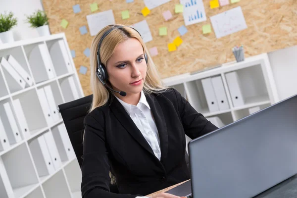 Geschäftsfrau in schwarzer Jacke und Kopfhörer mit Mikrofon am Computer. Büro im Hintergrund. Arbeitsbegriff. — Stockfoto