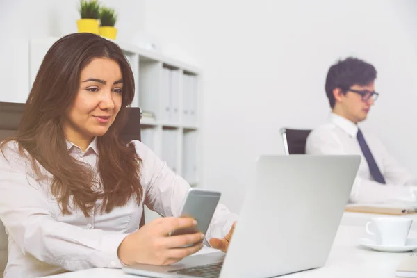 Задоволена бізнес-леді зі смартфоном в руці і бізнесменом, використовуючи ноутбук на офісному столі — стокове фото