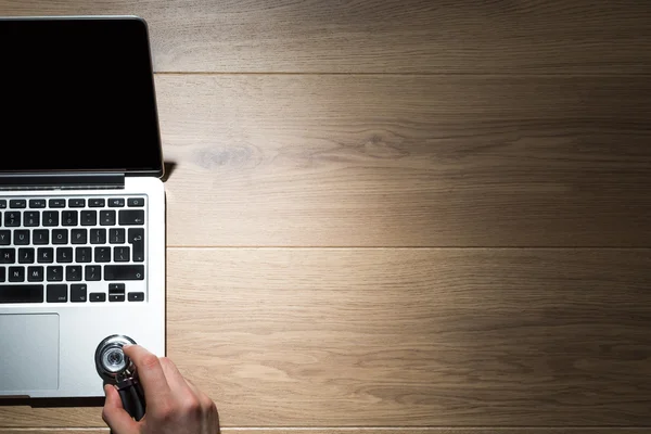 Vista superior del escritorio de madera con la mano masculina que escucha la computadora usando estetoscopio . — Foto de Stock